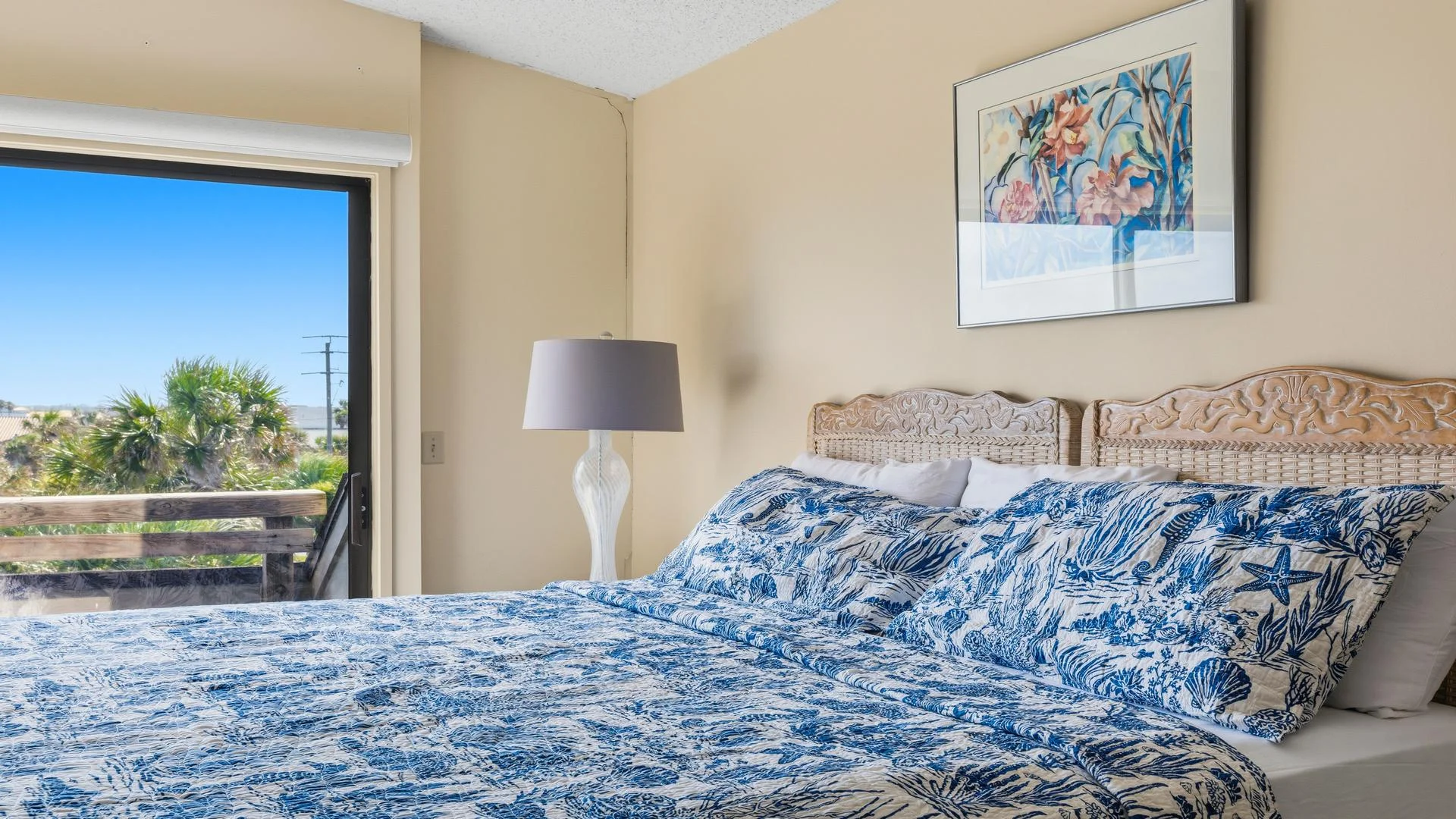 Glass sliding door opens to a wooden balcony railing with palm trees and blue sky beyond the bedroom.