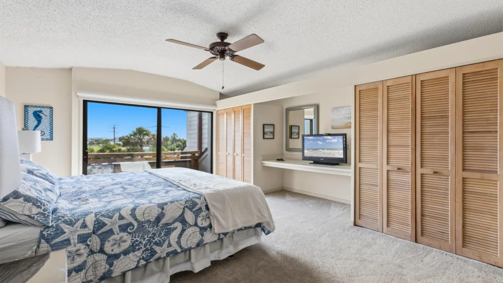 Bedroom with a queen bed covered in blue seashell-patterned bedding, sliding glass doors, and wood-paneled closets.