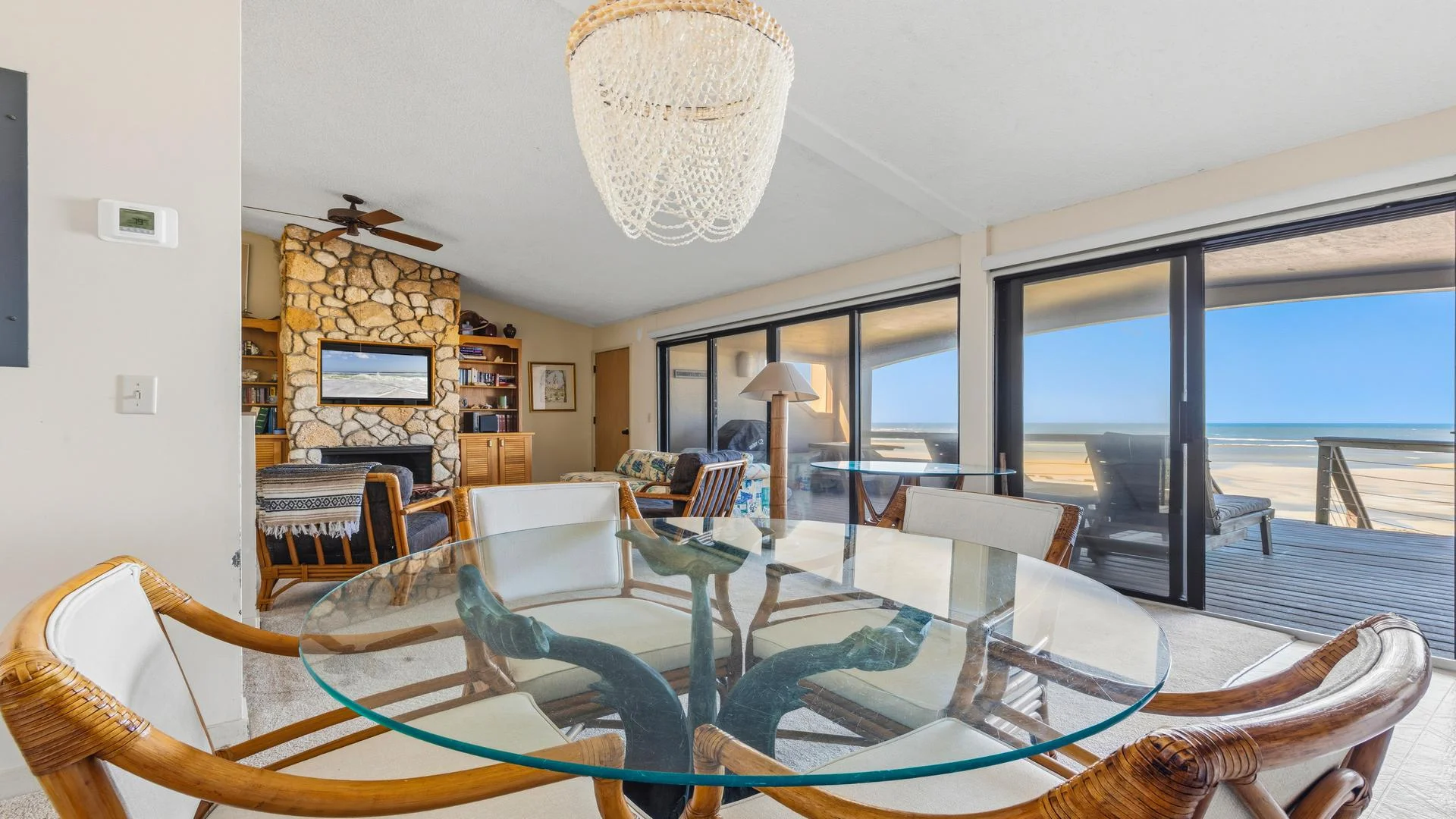 Glass-top dining table with rattan chairs faces sliding doors opening to oceanfront deck and sandy beach views.