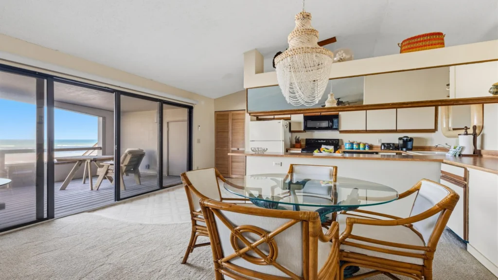 Dining area with glass-top table and rattan chairs beside sliding glass doors opening to an oceanfront balcony.