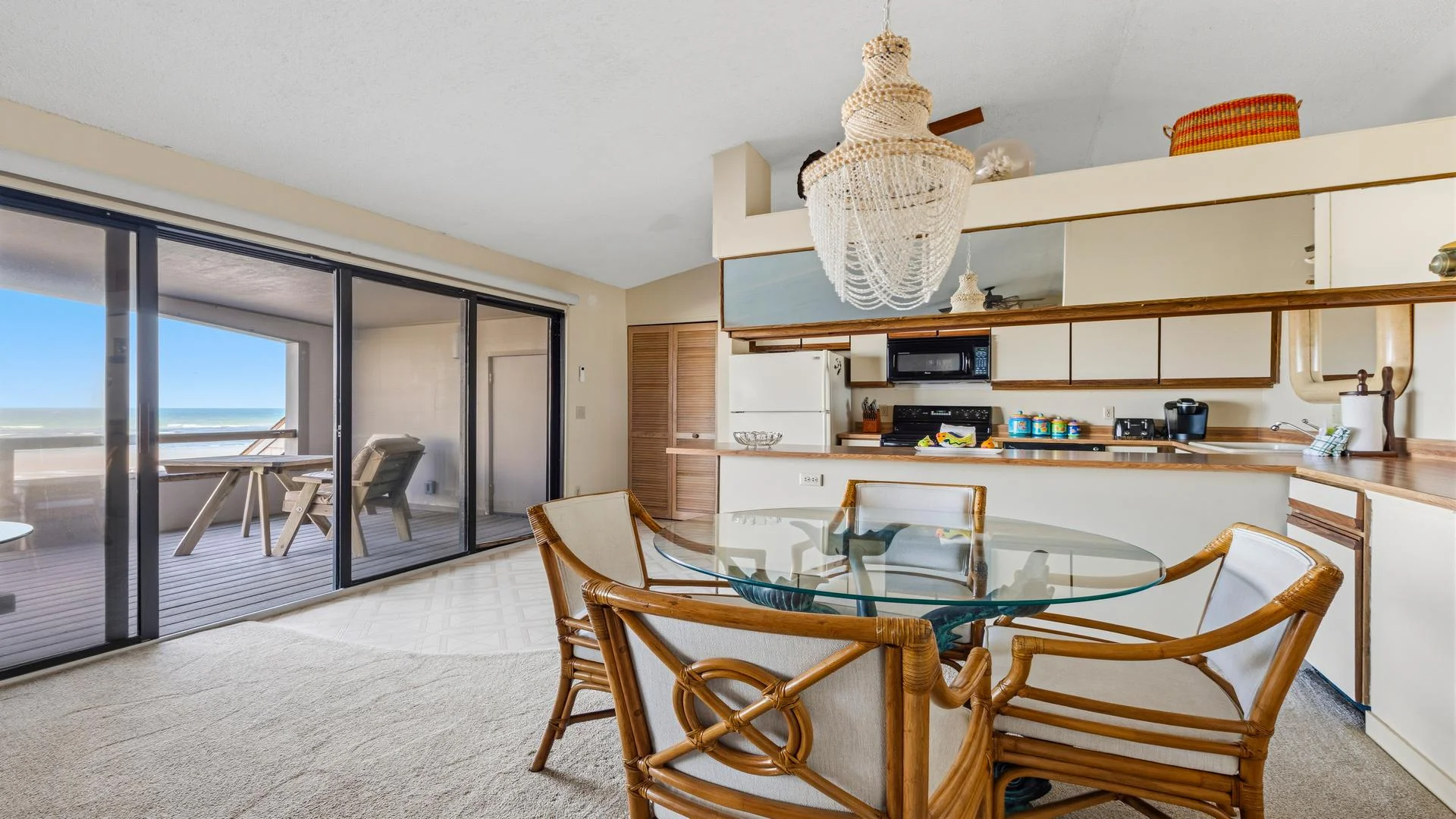 Dining area with glass-top table and rattan chairs beside sliding glass doors opening to an oceanfront balcony.