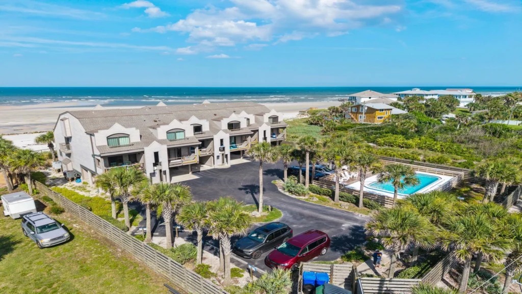 Three-story condo complex with asphalt parking, palm trees, fenced pool area, and oceanfront beach view.