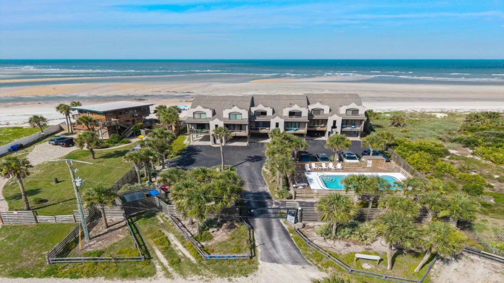 Aerial view of beachfront condos with gated entrance, palm trees, parking area, fenced communal pool, and sandy beach beyond.