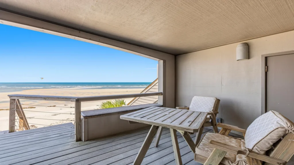 Covered balcony with weathered wood flooring, cushioned wooden chairs, and a cable-railed view of sandy beach and ocean.
