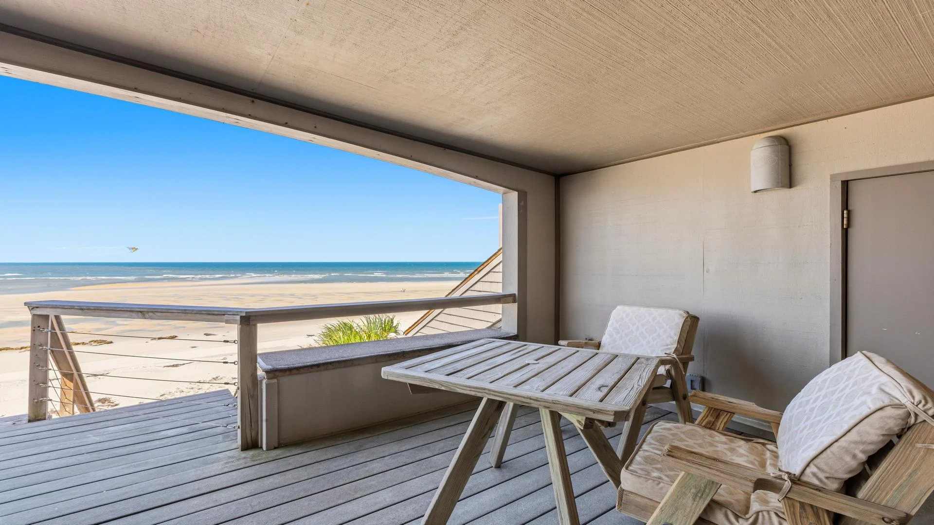 Covered balcony with weathered wood flooring, cushioned wooden chairs, and a cable-railed view of sandy beach and ocean.