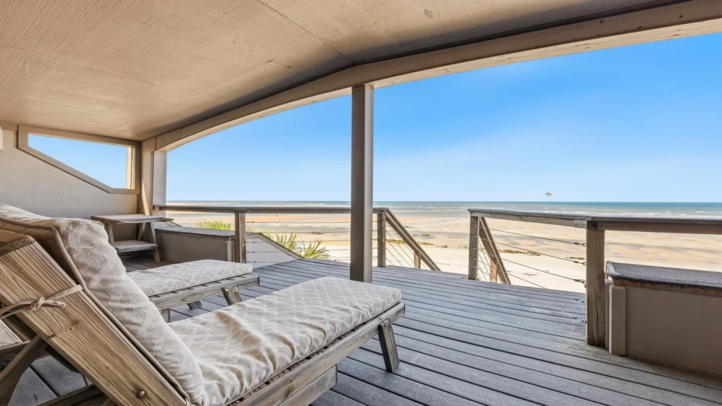 Two cushioned wooden chaise lounges sit on a covered deck overlooking the sandy beach and ocean.