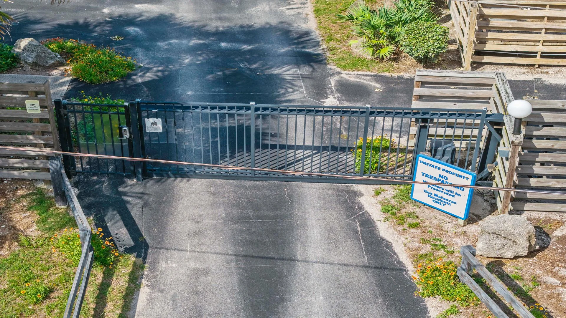 Black metal gate with vertical bars and a keypad entry secures the paved driveway with wooden fencing and greenery.