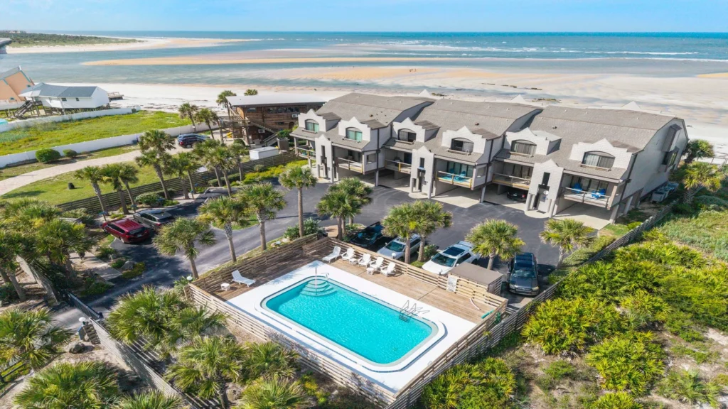 Air-conditioned condos with front balconies overlook a fenced pool area and white sandy beach beyond.