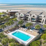 Air-conditioned condos with front balconies overlook a fenced pool area and white sandy beach beyond.