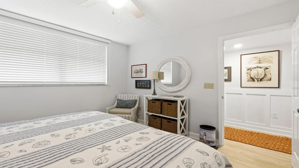 Guest bedroom with seashell-patterned bedding, woven wicker chair, white storage unit, and round wall mirror.