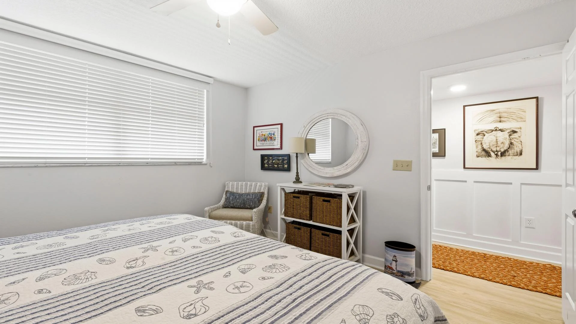 Guest bedroom with seashell-patterned bedding, woven wicker chair, white storage unit, and round wall mirror.