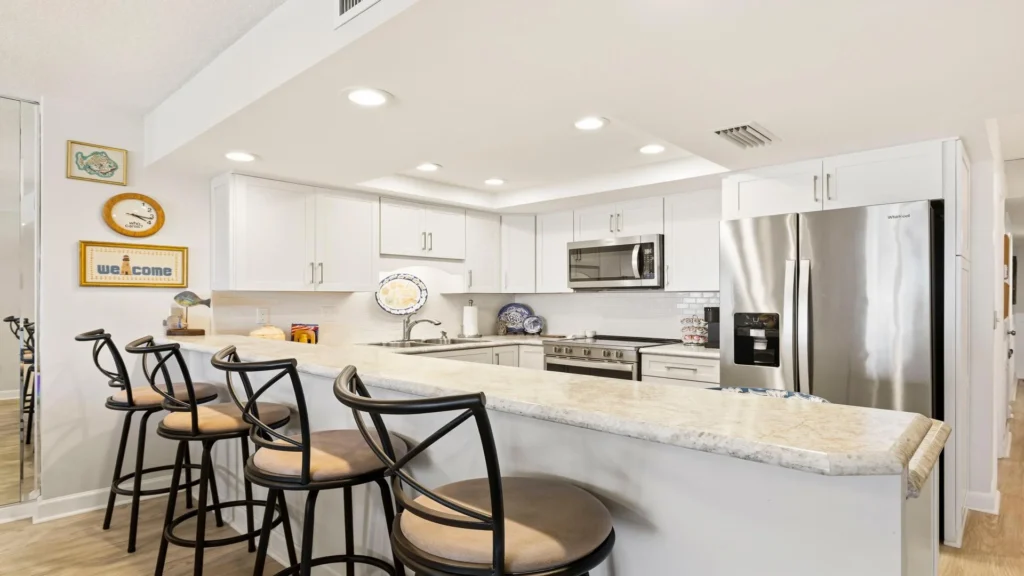 Marble countertop bar with four black metal stools faces white cabinetry and stainless steel appliances in the kitchen.