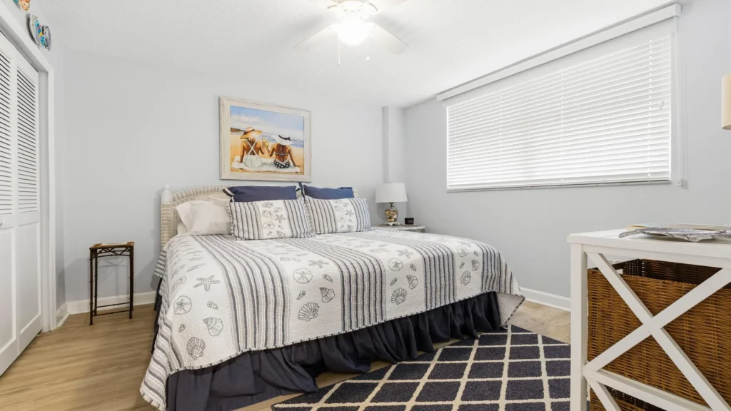 Bedroom with seashell-patterned bedding, wicker headboard, dark diamond rug, and white blinds over window.