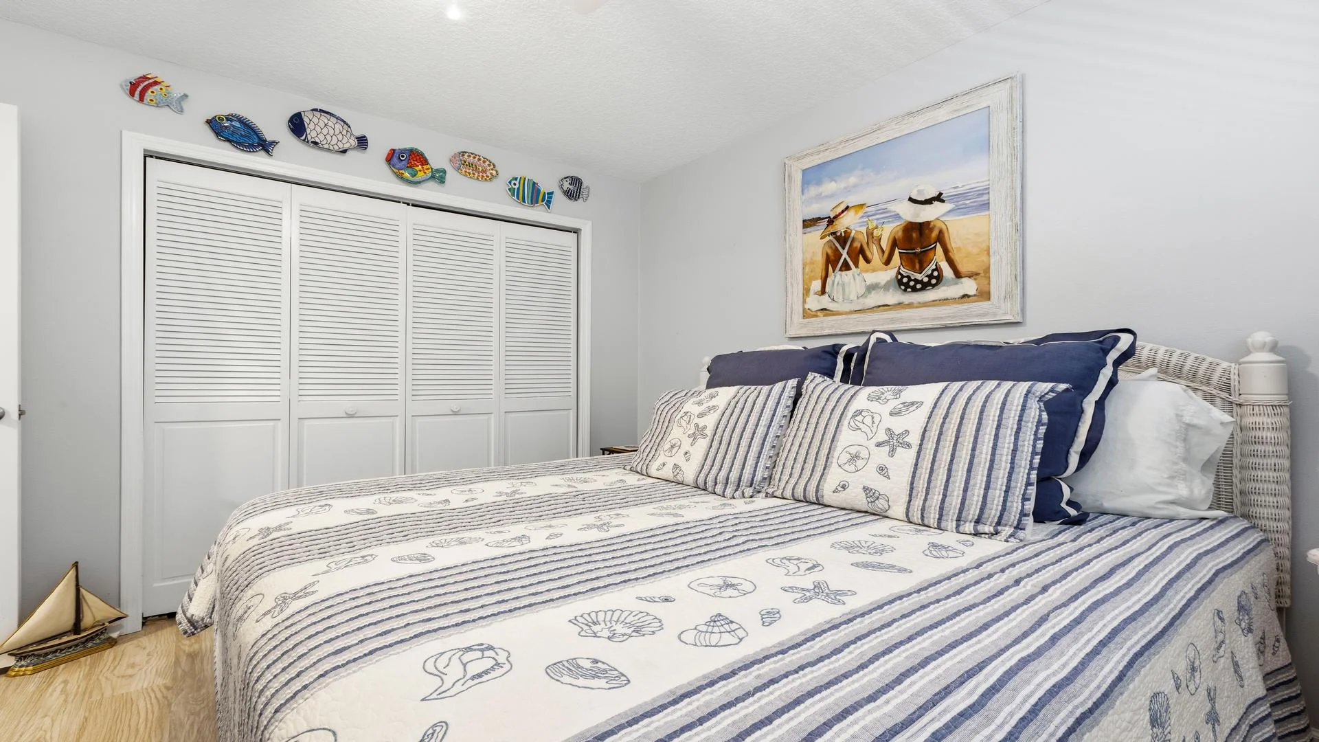 Bedroom with wicker headboard, striped seashell bedding, navy pillows, boat model, and colorful fish wall decor.