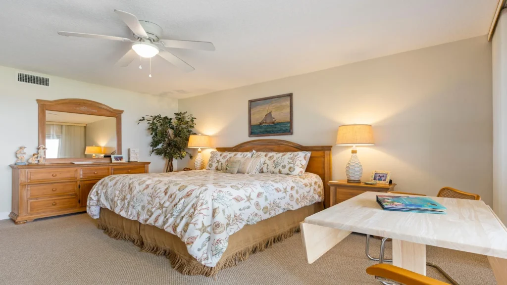 Master bedroom with seashell-patterned bedding, wooden dresser and nightstand, table lamp, and light wood desk with chair.