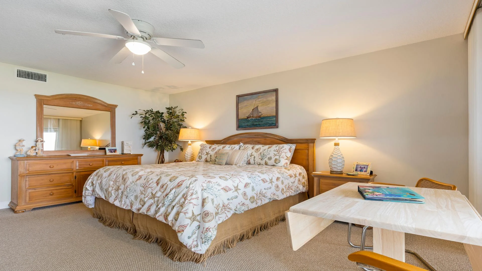 Master bedroom with seashell-patterned bedding, wooden dresser and nightstand, table lamp, and light wood desk with chair.