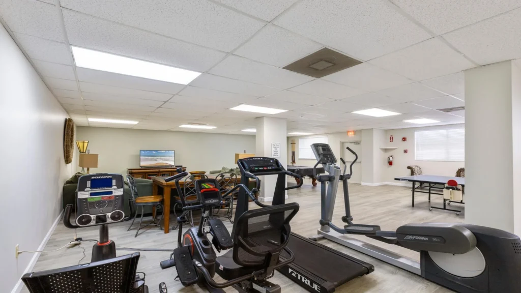 Fitness room equipped with recumbent bike, treadmill, elliptical, and a ping pong table beside cushioned seating.