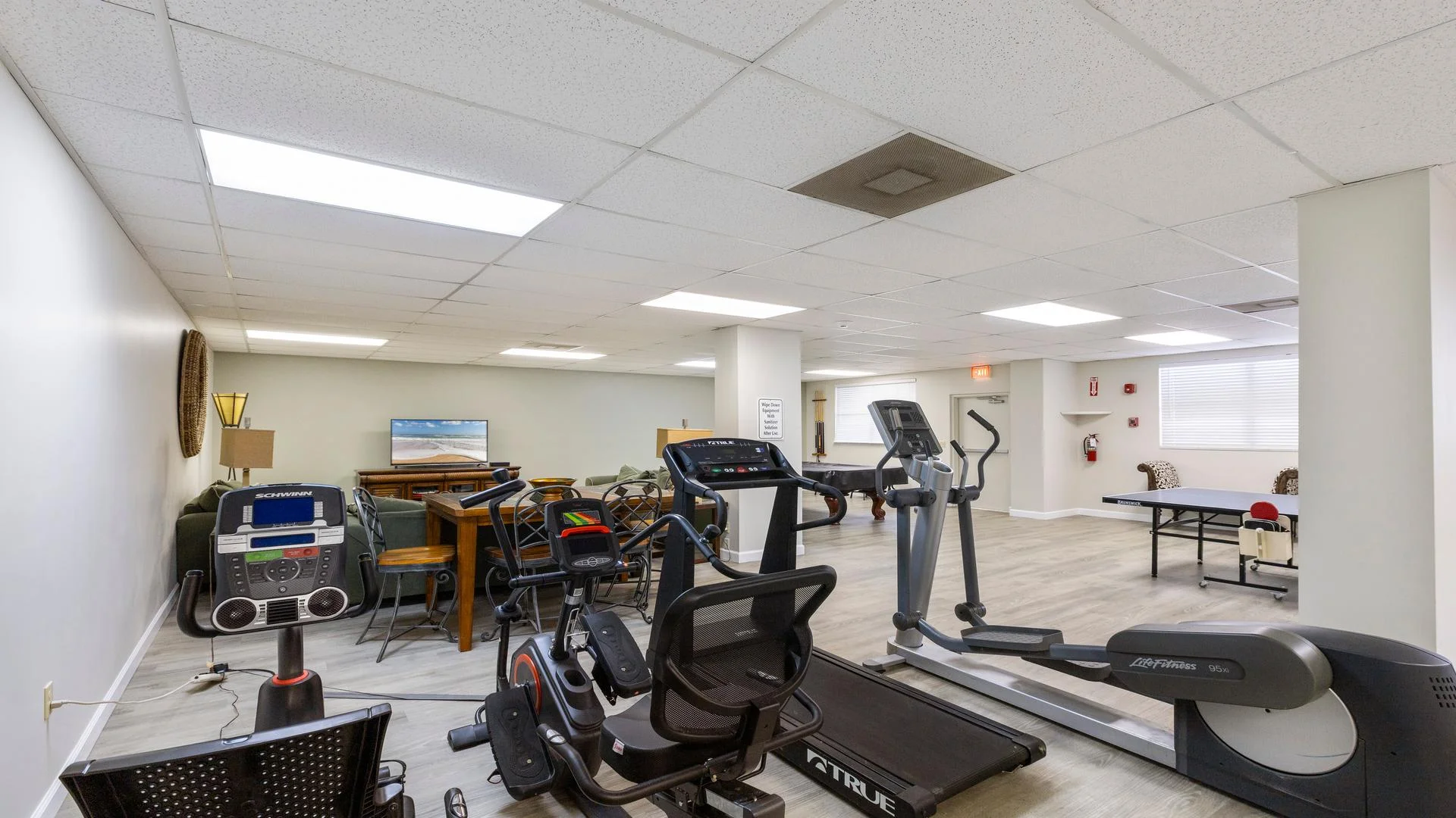 Fitness room equipped with recumbent bike, treadmill, elliptical, and a ping pong table beside cushioned seating.