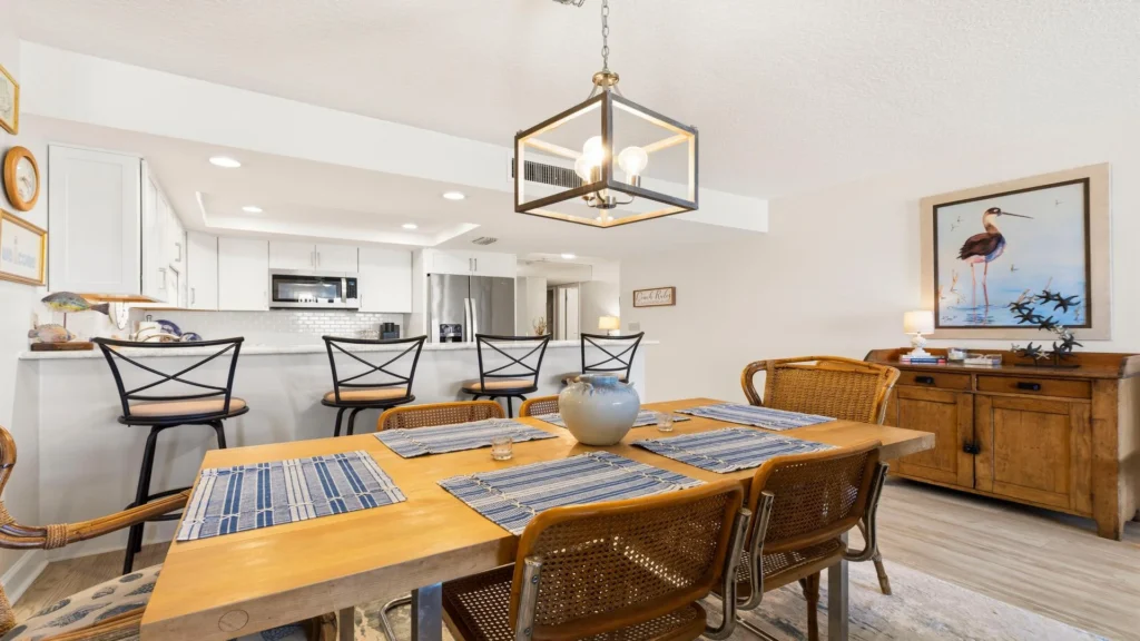 Dining area with woven rattan chairs, wooden sideboard, coastal bird artwork, and a cube pendant light overhead.