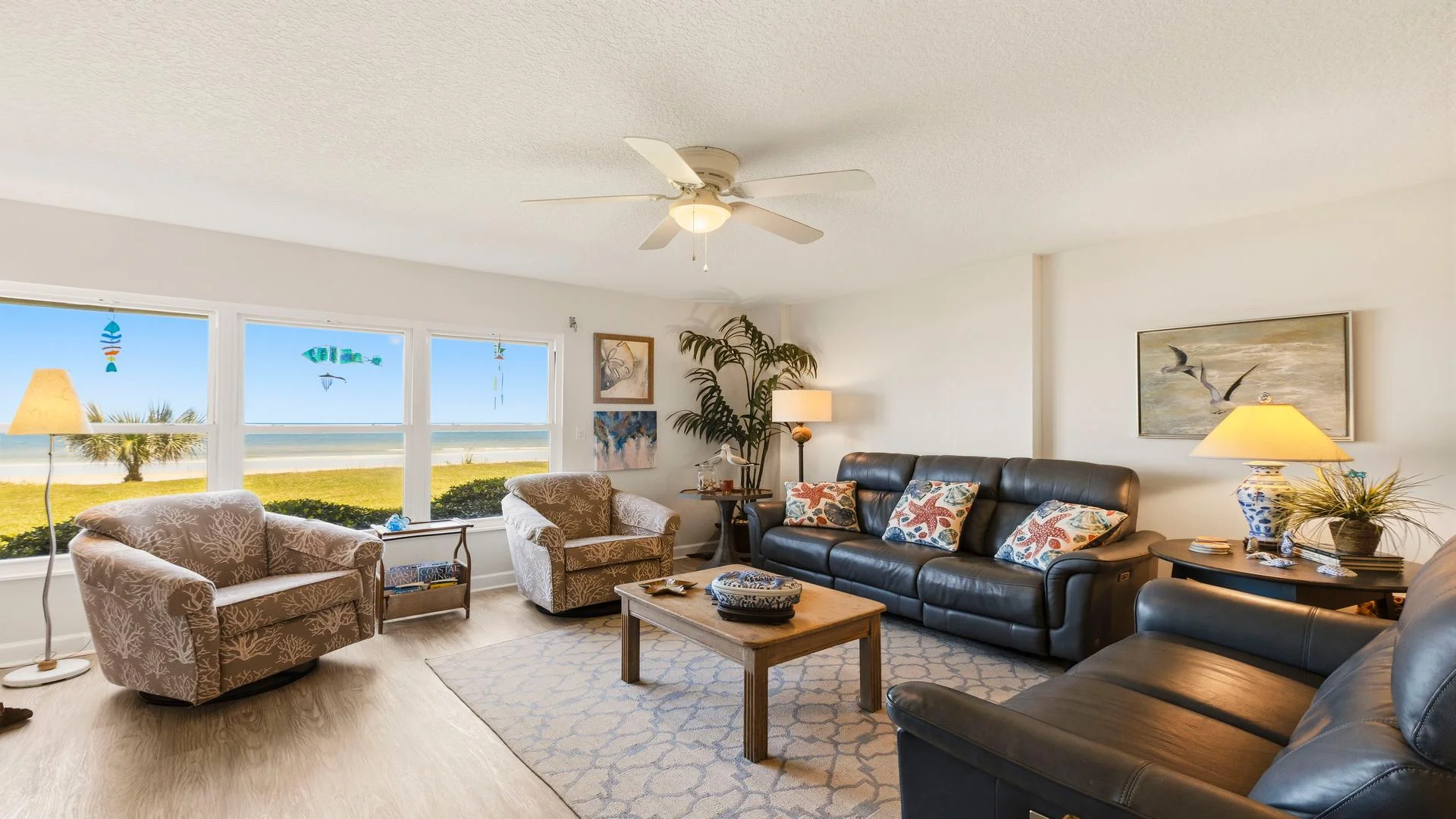 Living room with leather sofas, patterned armchairs, wooden coffee table, and ocean views through large windows.