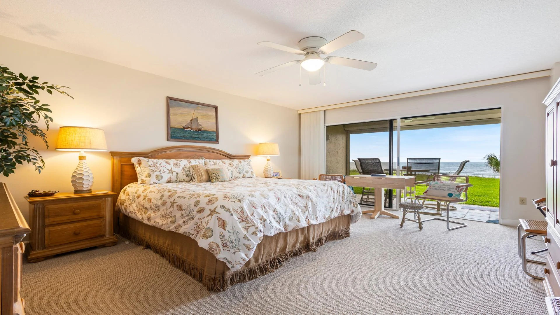 Sliding glass doors in the master bedroom open to a patio with outdoor seating and ocean views beyond green lawn.