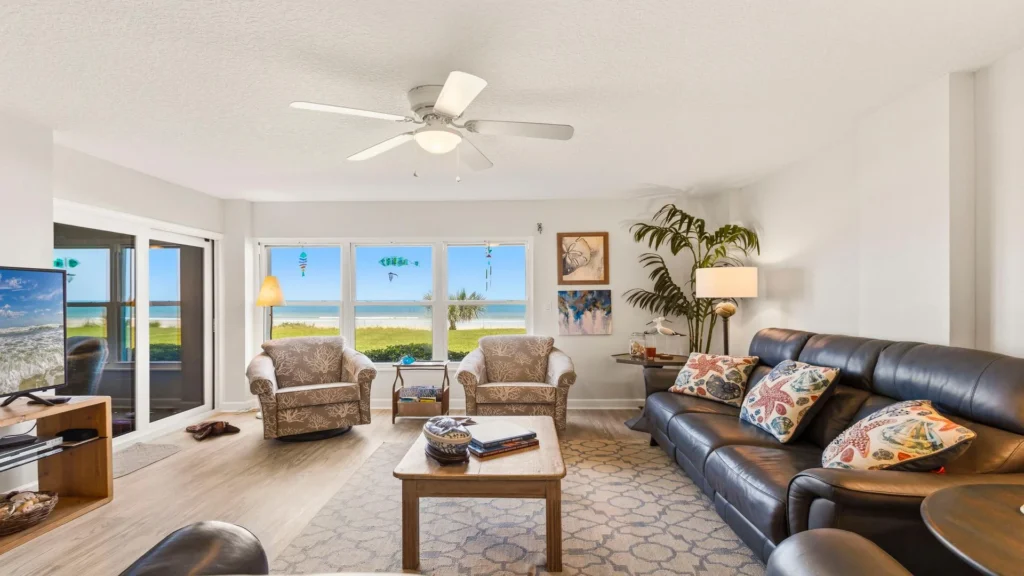 Living room with leather sofa, patterned armchairs, coastal pillows, wood coffee table, and ocean views through windows.