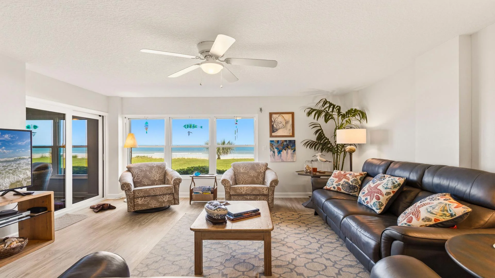 Living room with leather sofa, patterned armchairs, coastal pillows, wood coffee table, and ocean views through windows.