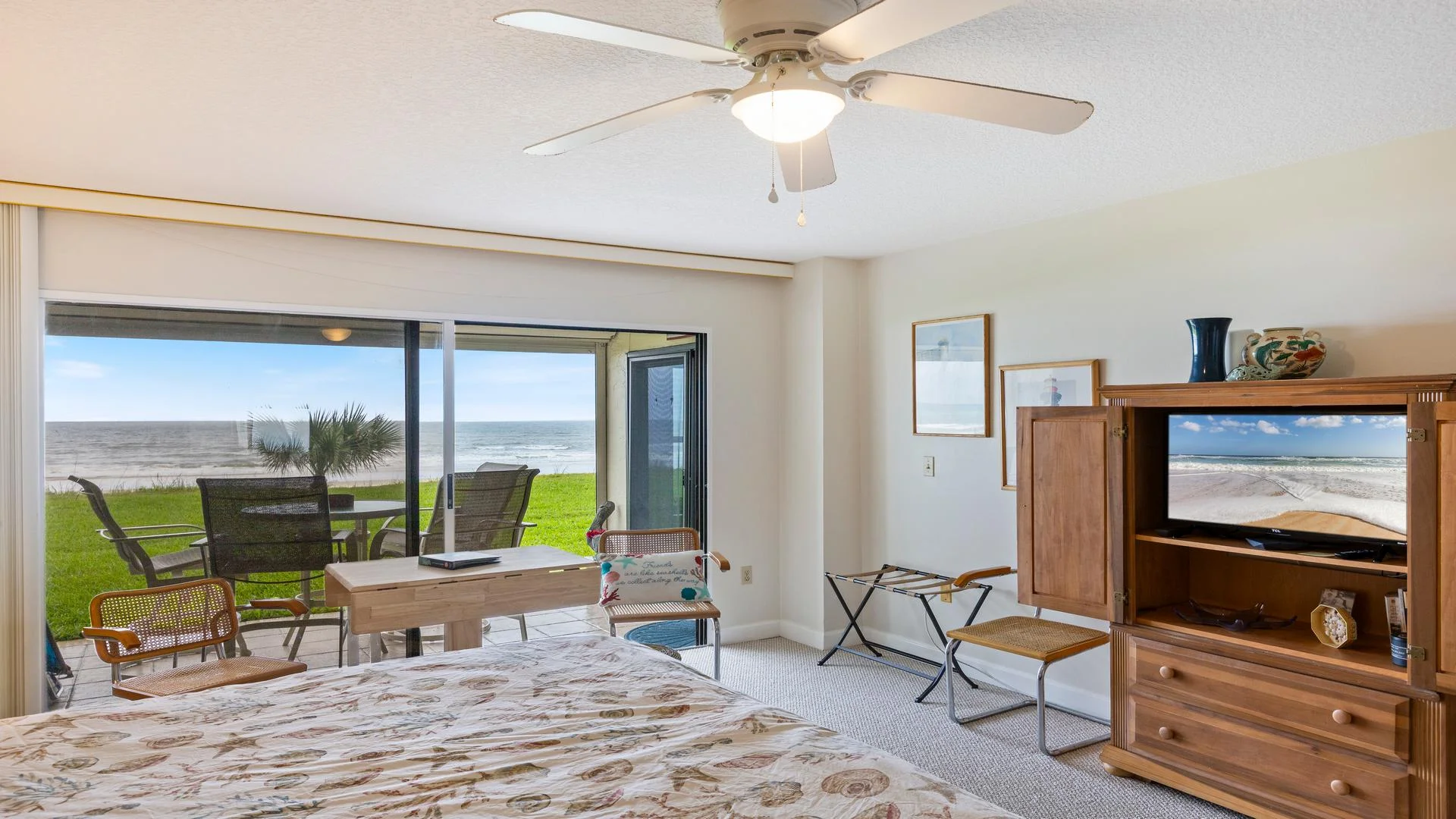 Oceanfront bedroom with seashell-patterned bedding, wood armoire TV cabinet, cane chairs, and sliding glass doors to patio.