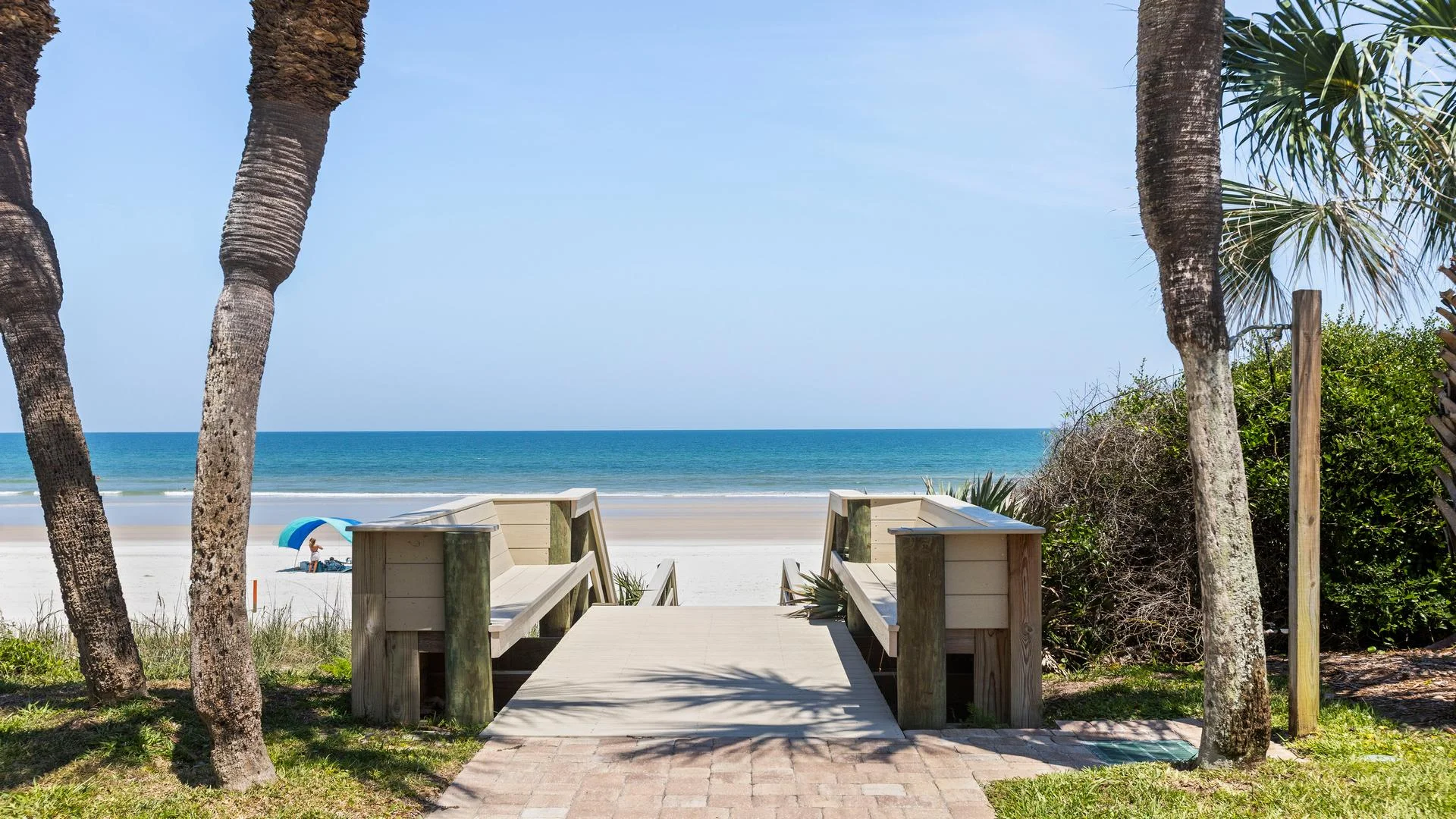Wooden boardwalk with built-in benches leads through palm trees to a sandy beach and blue ocean under clear sky.