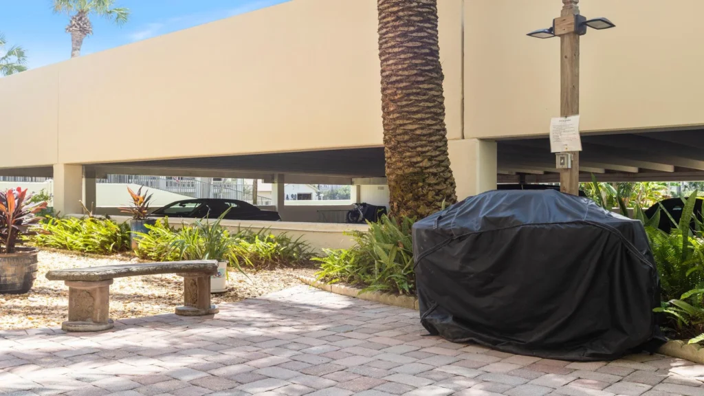Outdoor grilling area with a covered barbecue grill, stone bench, palm tree, and surrounding greenery.
