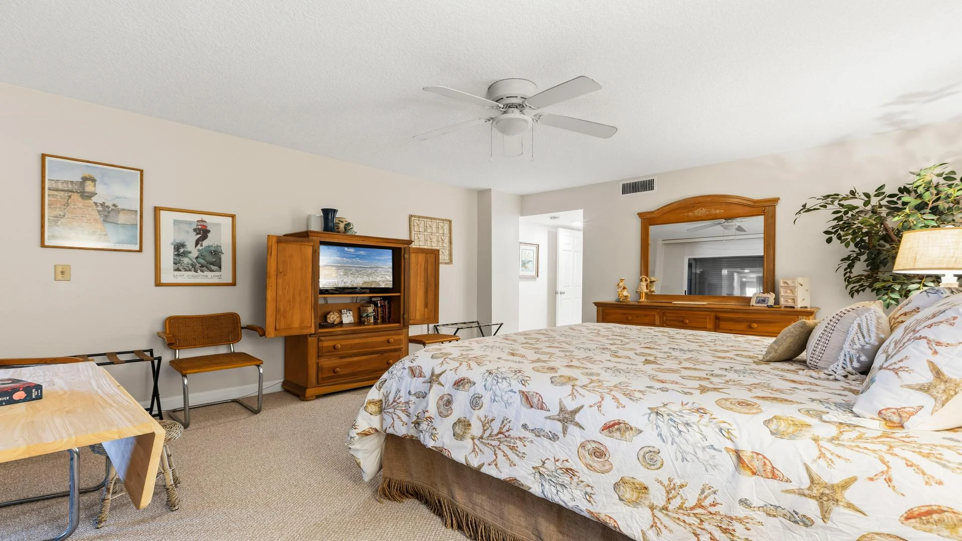 Light-filled bedroom with a seashell-patterned bedspread, wooden entertainment cabinet, and framed coastal prints.