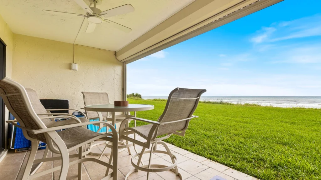 Covered patio with beige mesh chairs, round table, tiled floor, ceiling fan, and ocean views beyond green lawn.