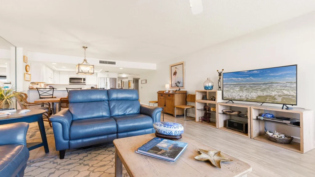 Living room with blue leather sofa, wood coffee table featuring coastal decor, and a TV on open shelving.