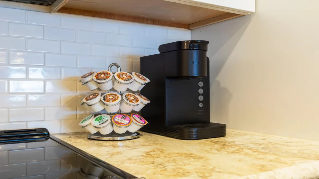Coffee station with black Keurig machine and rotating carousel of assorted coffee pods on granite countertop.