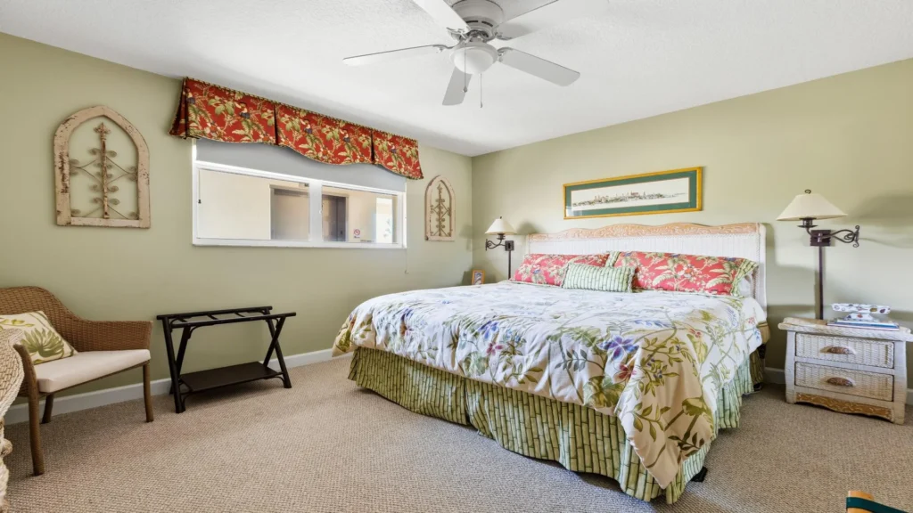 Room with muted green walls, a wicker headboard, floral bedding, beige carpet, and a red valance over a window.