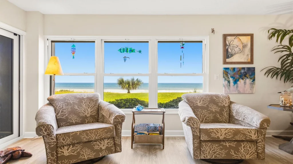Two patterned armchairs face wide windows framing an ocean view and coastal-themed wall art.