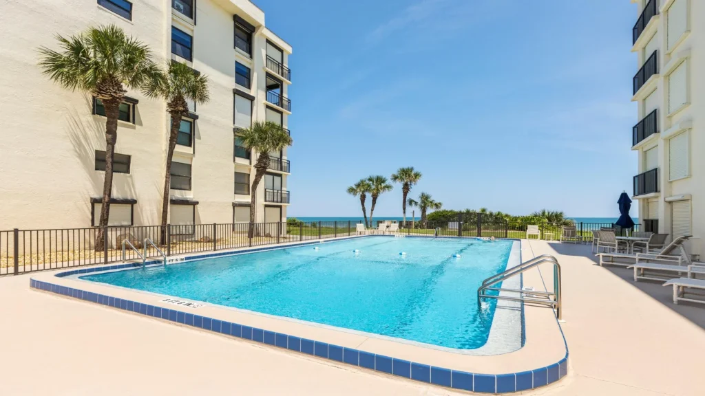 Outdoor swimming pool with blue tile trim, surrounded by lounge chairs and palm trees under clear skies.