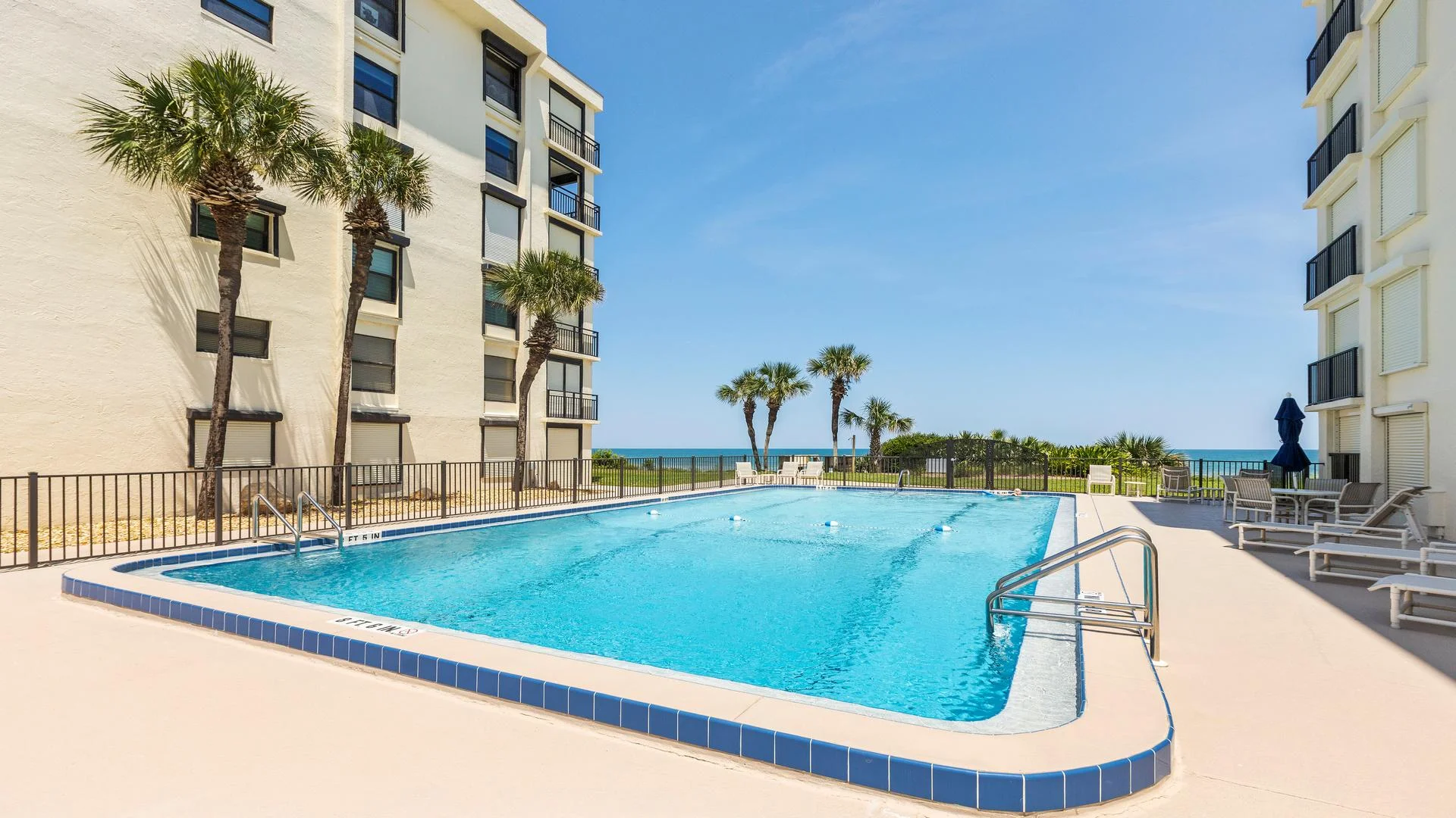 Outdoor swimming pool with blue tile trim, surrounded by lounge chairs and palm trees under clear skies.
