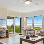 Living room with patterned armchairs, wooden coffee table, large windows, sliding glass door, and ocean views.