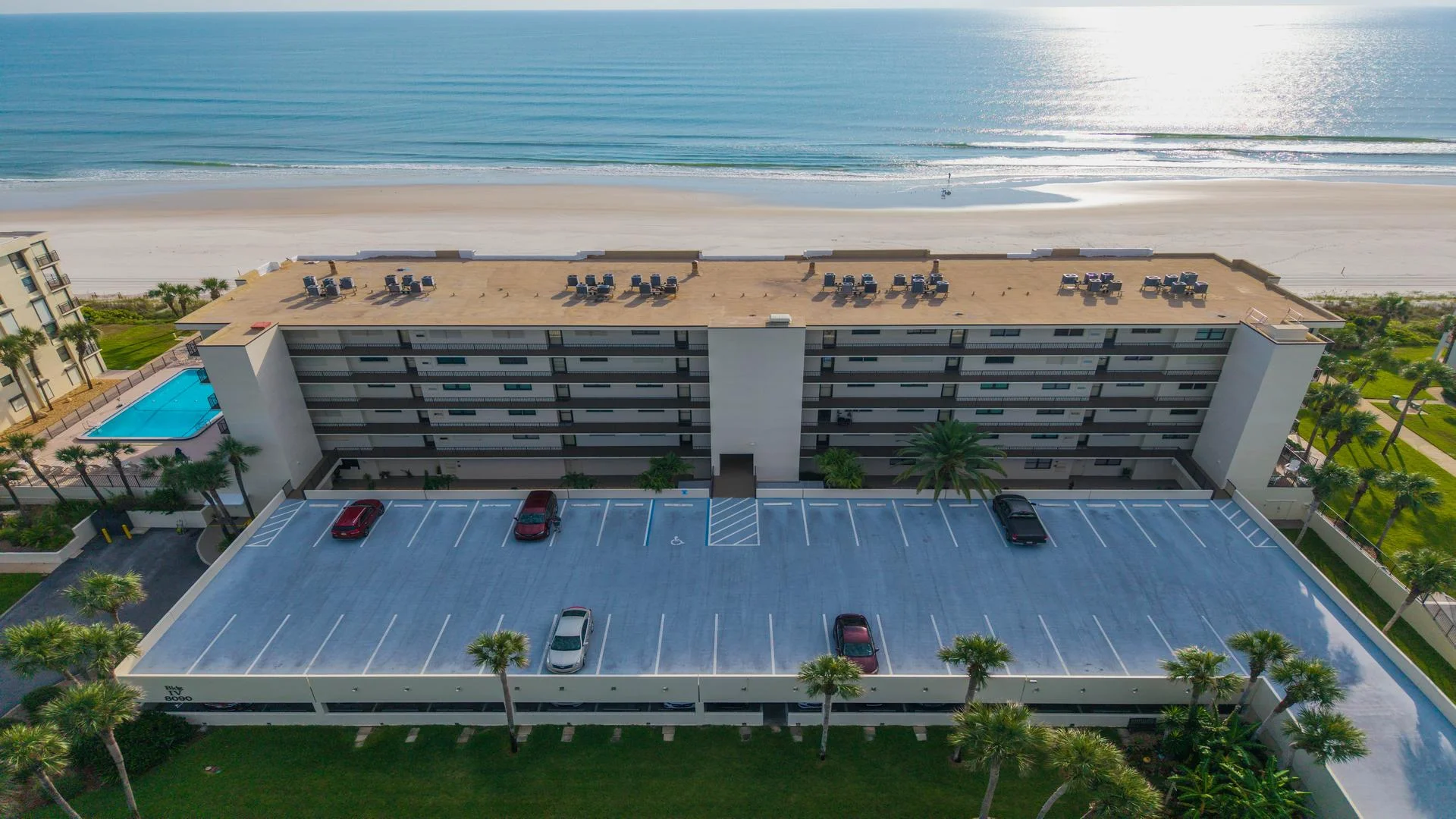 Aerial view of the condo building with rooftop seating, adjacent pool, and a parking garage facing the beach.