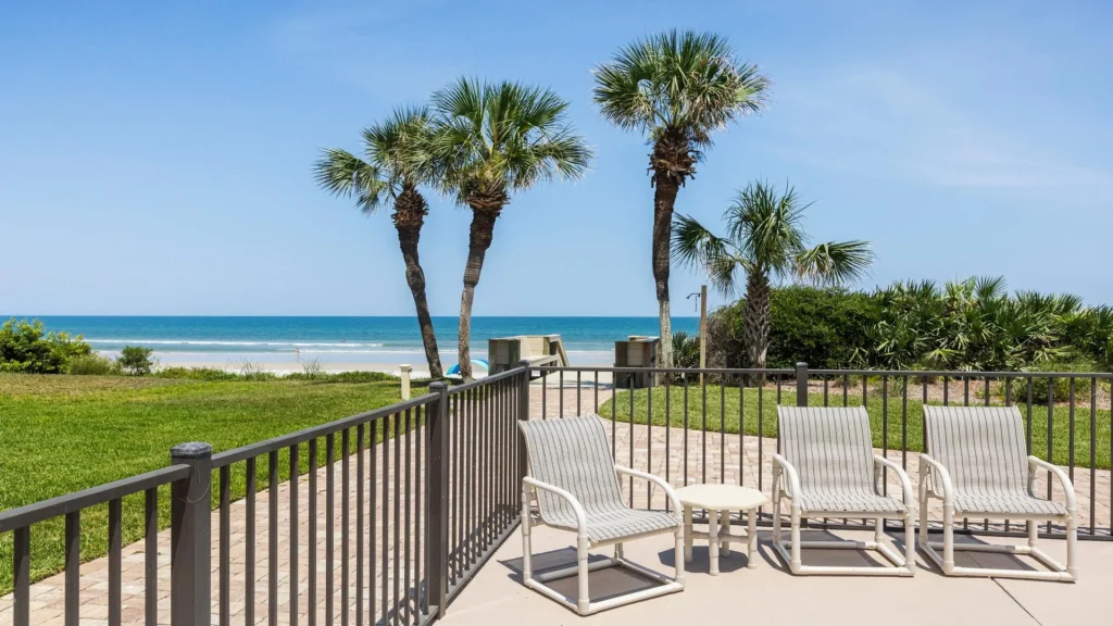 Patio seating with white metal chairs and table overlooks palm trees, sandy beach, and ocean horizon.