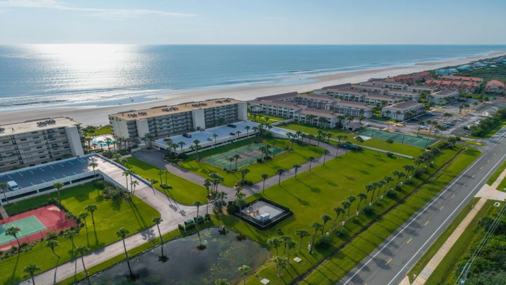 Aerial view of beachfront condos with tennis courts, palm-lined streets, parking structures, and ocean waves.