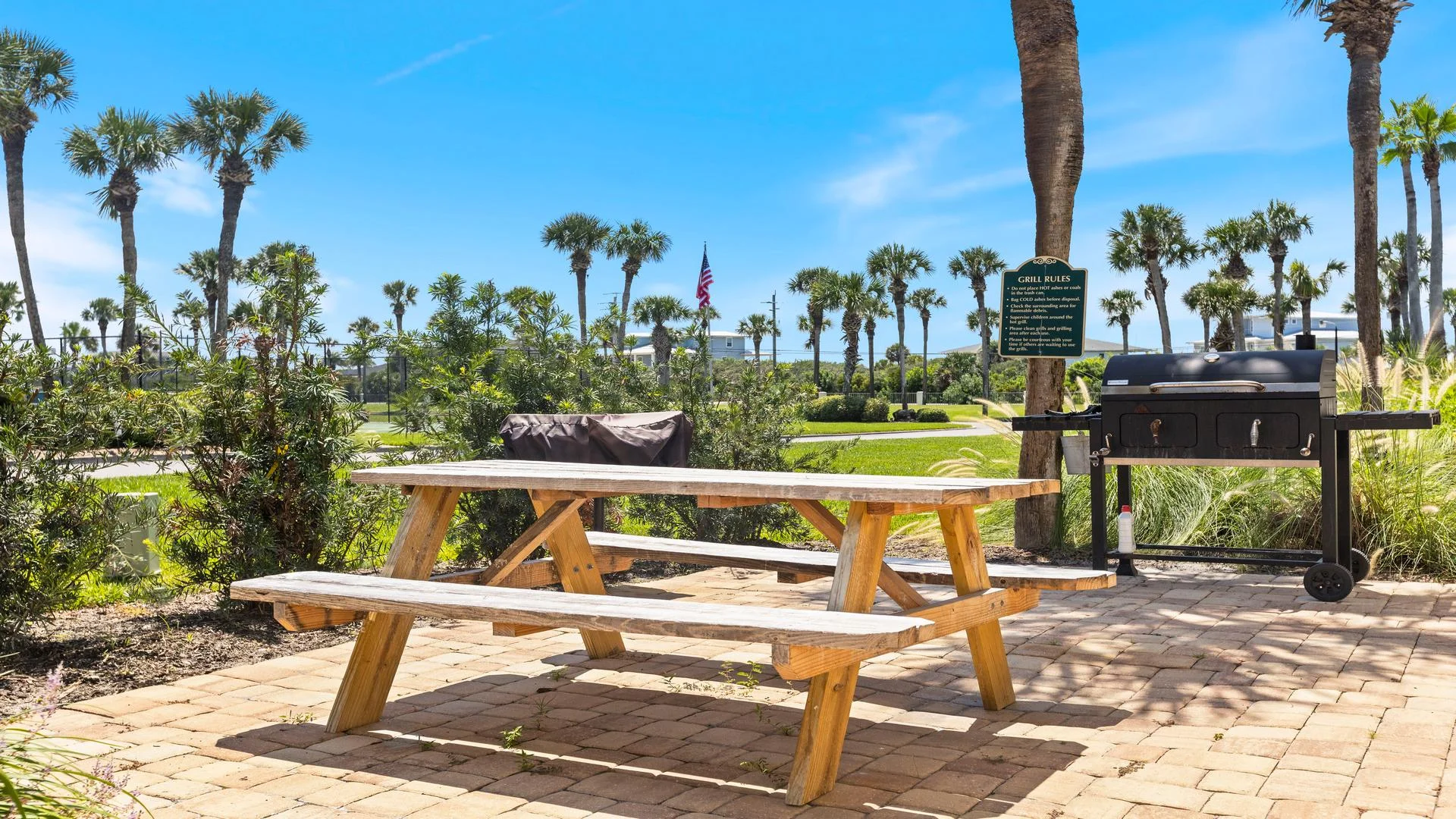 Outdoor grilling area with a charcoal grill, wooden picnic table, and palm trees under clear blue sky.