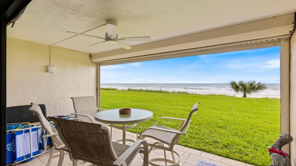 Covered patio with glass-top table, four mesh chairs, ceiling fan, and oceanfront lawn view.