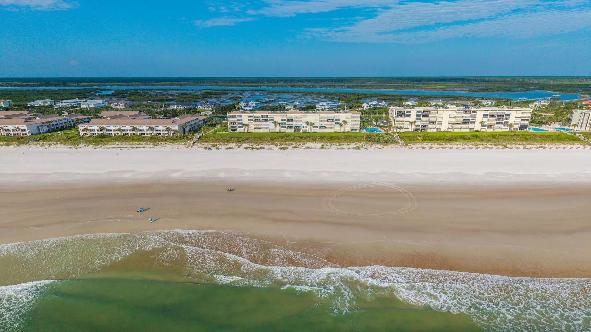 Wide aerial view of beachfront condos with sandy shore, gentle waves, and marshland in the distance under a blue sky.