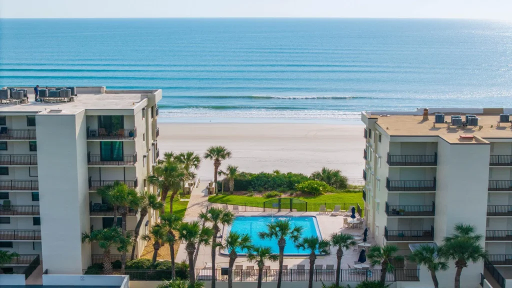 Aerial view of the pool area framed by palm trees between two white condo buildings, with sandy beach and ocean beyond.