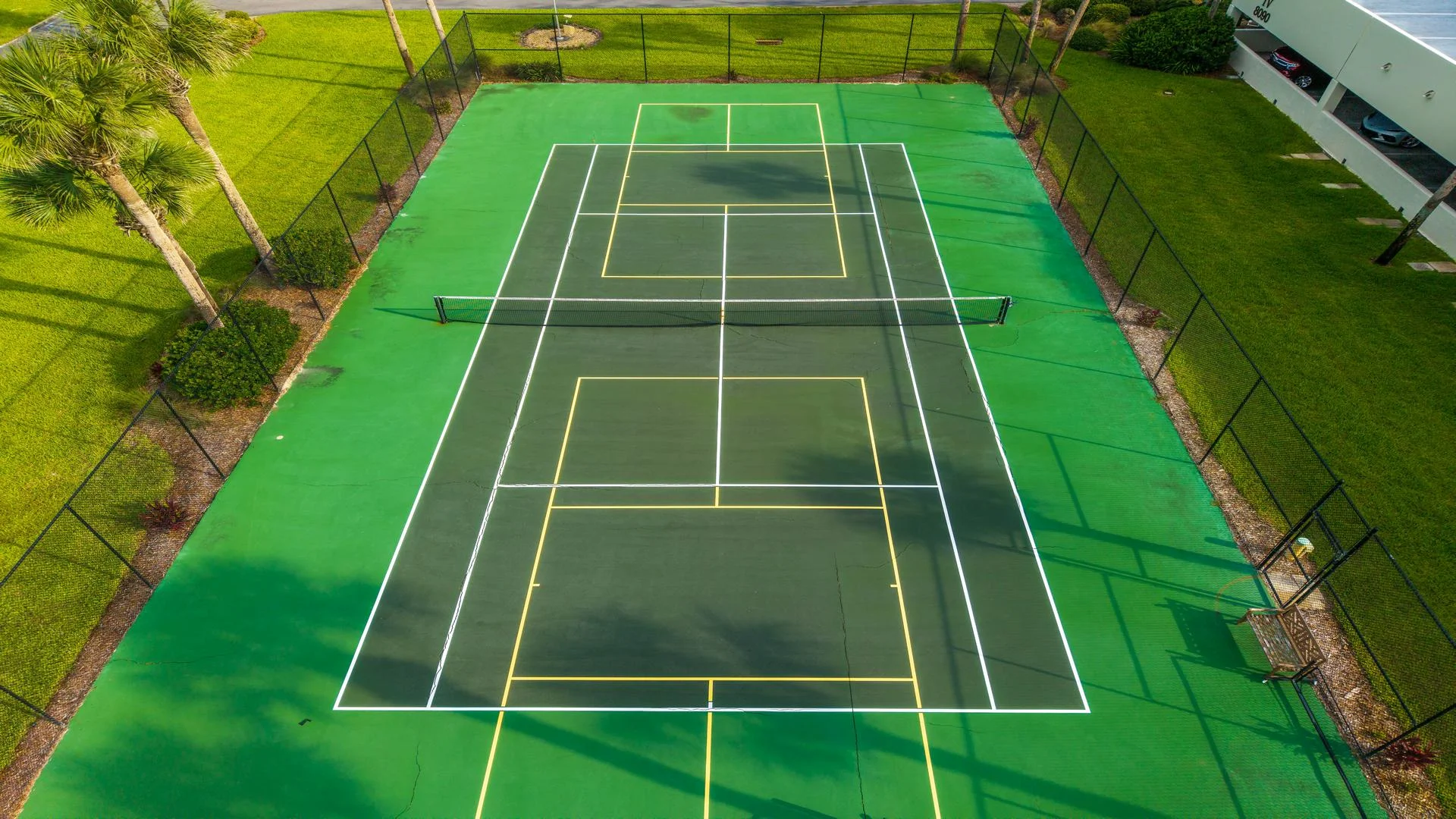 Outdoor tennis court with green and dark green surface, white and yellow boundary lines, black net, and surrounding fence.