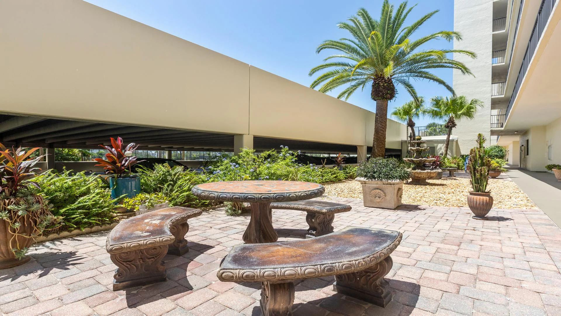 Outdoor seating area with curved stone benches and table, surrounded by potted plants and palm trees.