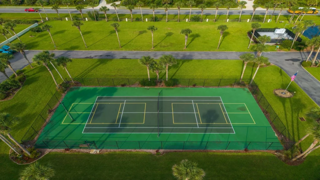 Aerial view of the fenced tennis court with green surface, surrounded by palm trees and manicured grass.