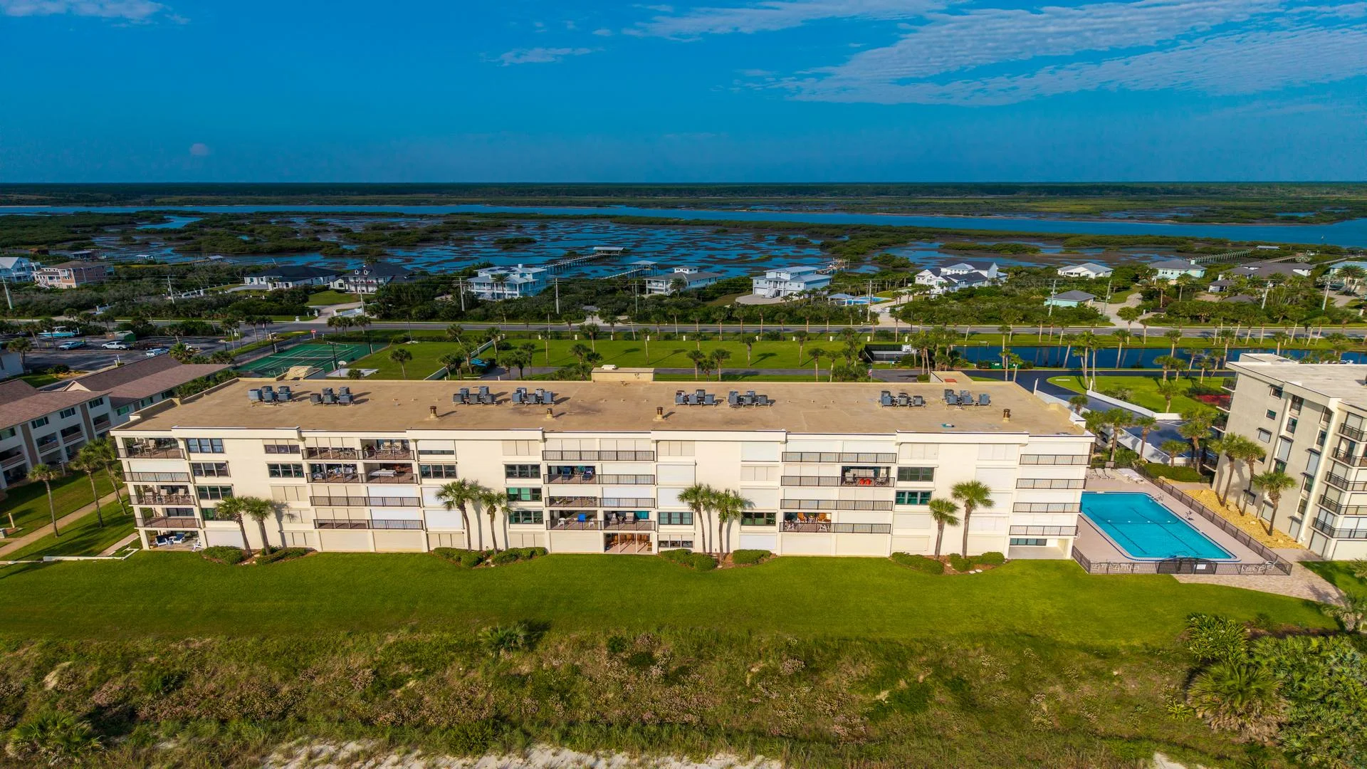 Exterior view of a low-rise condo building with balconies, palm trees, a swimming pool, and marshland beyond.
